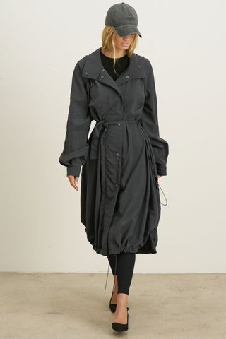 Full body front view of model posing in the jet nylon City Trench coat with oversized silhouette, structured collar, attached hood, self-tie belt and adjustable sweep
