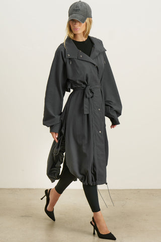 Full body front view of model posing in the jet nylon City Trench coat with oversized silhouette, structured collar, attached hood, self-tie belt and adjustable sweep