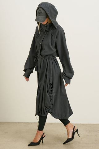 Full body side view of model posing in the jet nylon City Trench coat with oversized silhouette, structured collar, attached hood, self-tie belt and adjustable sweep