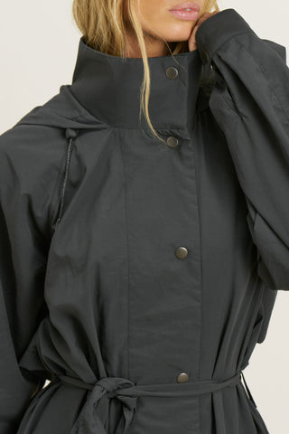 Close up detail front view of model posing in the jet nylon City Trench coat with oversized silhouette, structured collar, attached hood, self-tie belt and adjustable sweep