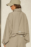 Back view of model posing in the oversized desert sage nylon Classic Windbreaker Jacket with a collar, zip-away hood and reflective corded details at the hem, sleeves and neck for cinching.