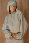 Front view of model posing in the oversized desert sage nylon Classic Windbreaker Jacket with a collar, zip-away hood and reflective corded details at the hem, sleeves and neck for cinching.