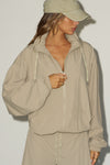 Front view of model posing in the oversized desert sage nylon Classic Windbreaker Jacket with a collar, zip-away hood and reflective corded details at the hem, sleeves and neck for cinching.