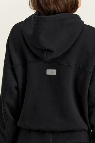 Close up back view of model posing outside in the relaxed fit black french terry Classic Zip Hoodie with a full length front zip closure, side pockets and thumbholes in the fitted cuffs