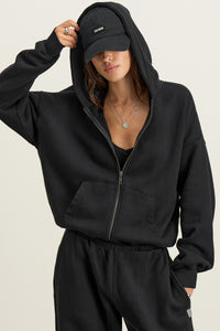 Front view of model posing outside in the relaxed fit black french terry Classic Zip Hoodie with a full length front zip closure, side pockets and thumbholes in the fitted cuffs