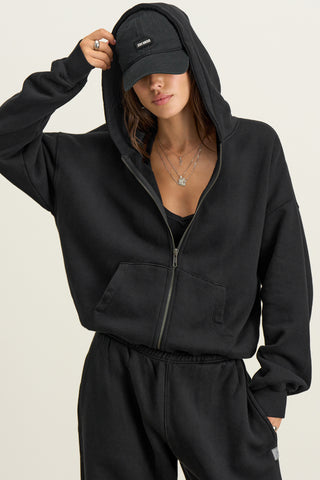 Front view of model posing outside in the relaxed fit black french terry Classic Zip Hoodie with a full length front zip closure, side pockets and thumbholes in the fitted cuffs