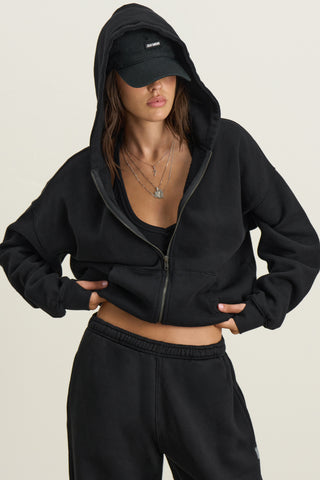 Front view of model posing outside in the relaxed fit black french terry Classic Zip Hoodie with a full length front zip closure, side pockets and thumbholes in the fitted cuffs