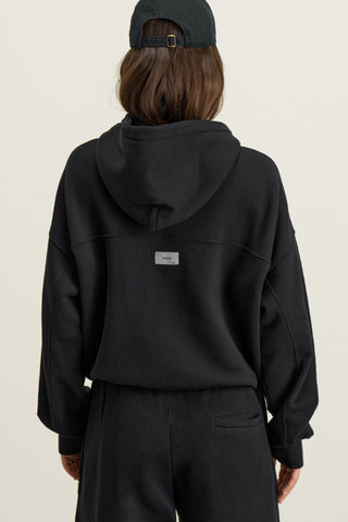 Back view of model posing outside in the relaxed fit black french terry Classic Zip Hoodie with a full length front zip closure, side pockets and thumbholes in the fitted cuffs