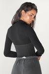 Back view of model posing in the fitted stretchy vintage black flexrib Corset Long Sleeve top with a mock neckline, modal top half and ribbed bottom half with corset-style seaming at the bodice