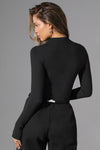 Back view of model posing in the fitted sueded onyx Cut Out Long Sleeve Top with front cutout details, three front adjustable clasps and thumbholes in the sleeve cuffs