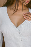 Close up detail front view of model posing in the soft and fitted pearl grey luxe knit Asymmetrical Button Mini Dress with long sleeves,  a v-neckline and asymmetrical button-front details. 