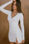 Front view of model posing in the soft and fitted pearl grey luxe knit Asymmetrical Button Mini Dress with long sleeves,  a v-neckline and asymmetrical button-front details. 