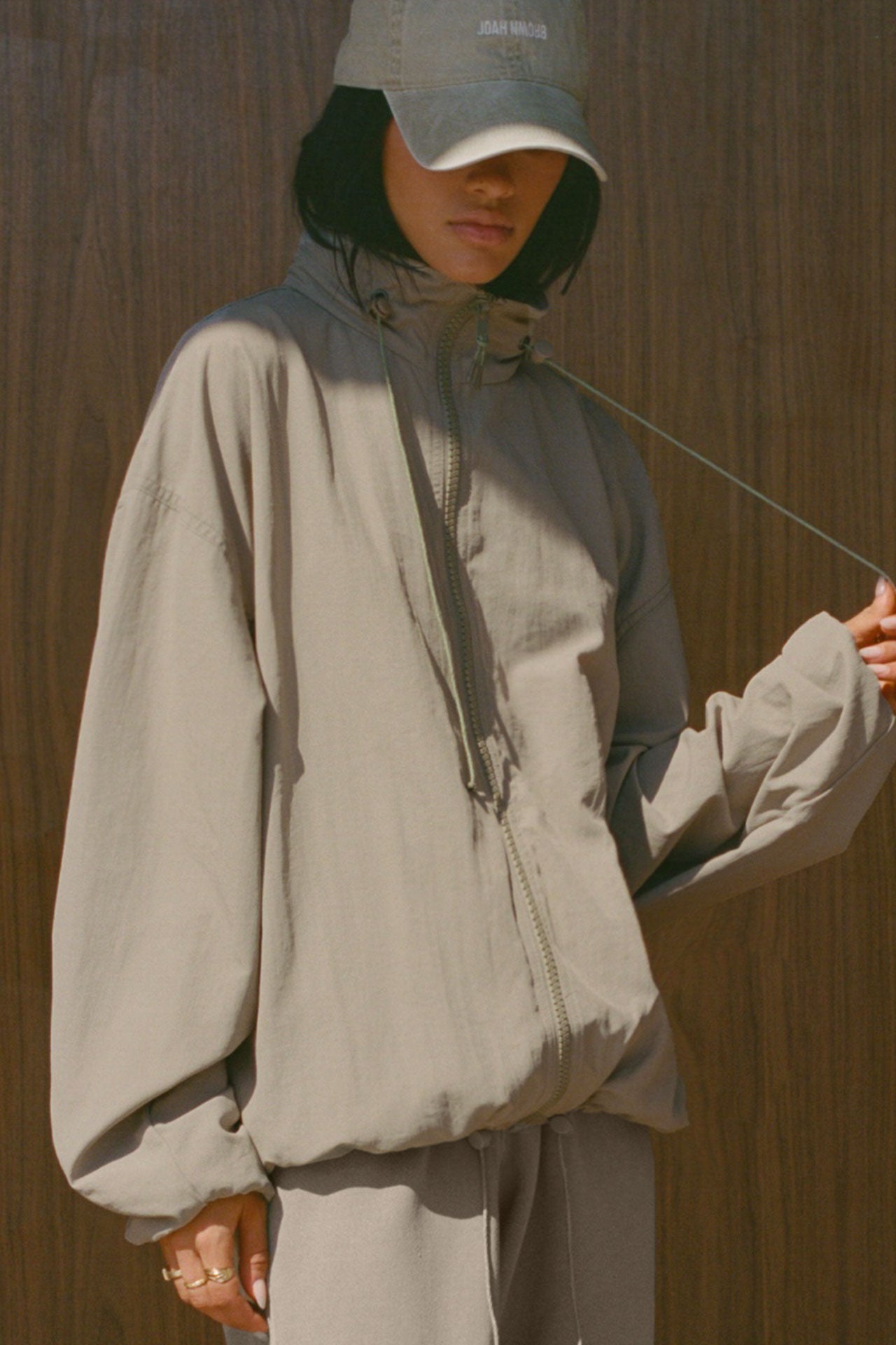 Front view of model posing in the oversized desert sage nylon Classic Windbreaker Jacket with a collar, zip-away hood and reflective corded details at the hem, sleeves and neck for cinching.