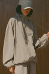 Front view of model posing in the oversized desert sage nylon Classic Windbreaker Jacket with a collar, zip-away hood and reflective corded details at the hem, sleeves and neck for cinching.