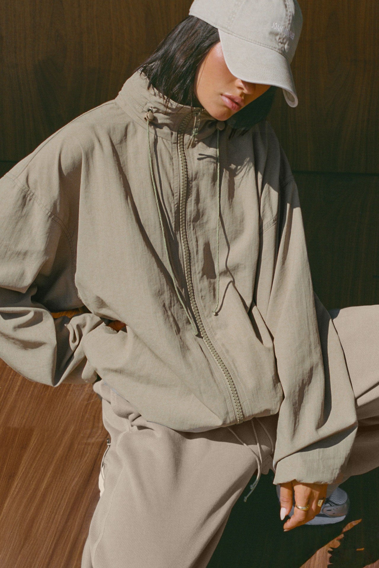 Front view of model posing in the oversized desert sage nylon Classic Windbreaker Jacket with a collar, zip-away hood and reflective corded details at the hem, sleeves and neck for cinching.