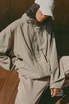 Front view of model posing in the oversized desert sage nylon Classic Windbreaker Jacket with a collar, zip-away hood and reflective corded details at the hem, sleeves and neck for cinching.