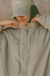 Close up detail front view of model posing in the oversized desert sage nylon Classic Windbreaker Jacket with a collar, zip-away hood and reflective corded details at the hem, sleeves and neck for cinching.