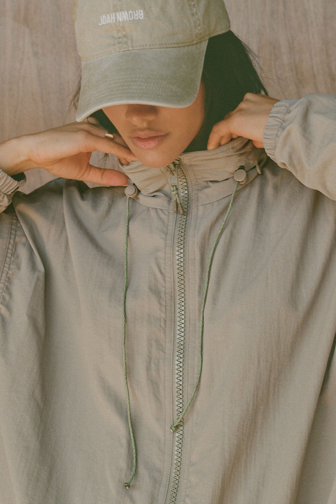 Close up detail front view of model posing in the oversized desert sage nylon Classic Windbreaker Jacket with a collar, zip-away hood and reflective corded details at the hem, sleeves and neck for cinching.