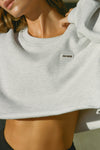 Close up detail front view of model posing in the loose fit cropped pearl grey french terry Cut Off Sweatshirt with oversized long sleeves, a raw cut hem and a white patch with a black joah brown logo on it sewn on the upper left chest