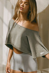 Front view of model posing in the relaxed fit cropped haze cotton Off Shoulder Crop Tee with a JOAH BROWN logo patch on the left sleeve and a wide scoop neckline that can be worn off the shoulders