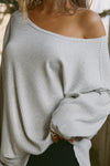 Close up detail front view of model posing in the relaxed fit pearl grey luxe knit Off Shoulder Tunic with a wide off-the-shoulder neckline and asymmetrical hem