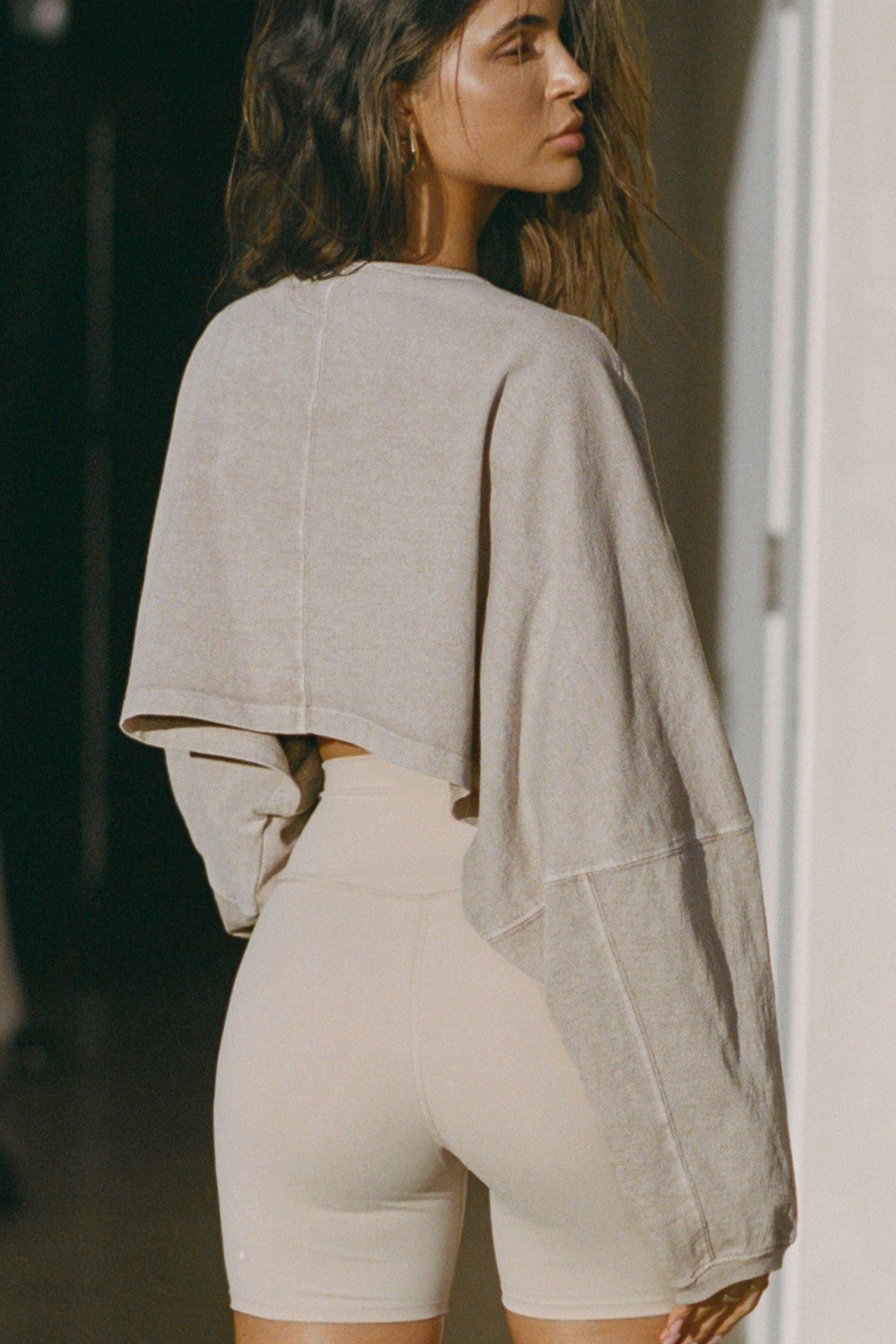 Back view of model posing in the cropped relaxed fit sandstone cotton Slouchy Crop Long Sleeve top with a crew neckline, dropped shoulders and oversized sleeves