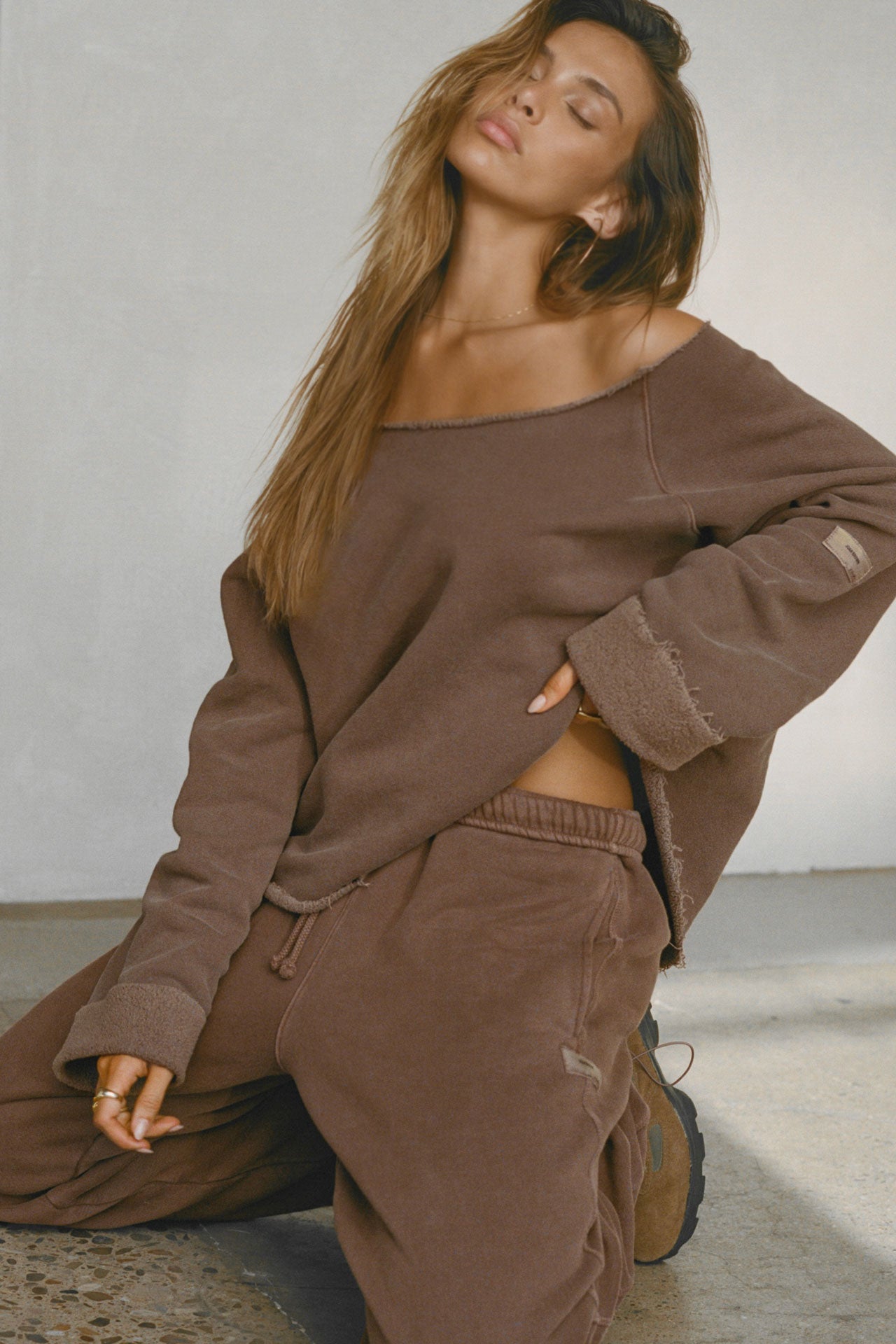 Front view of model kneeling on the floor posing in the oversized cedar french terry Slouchy Pullover Sweatshirt with a wide, off-the-shoulder neckline and a JOAH BROWN logo patch on the right sleeve