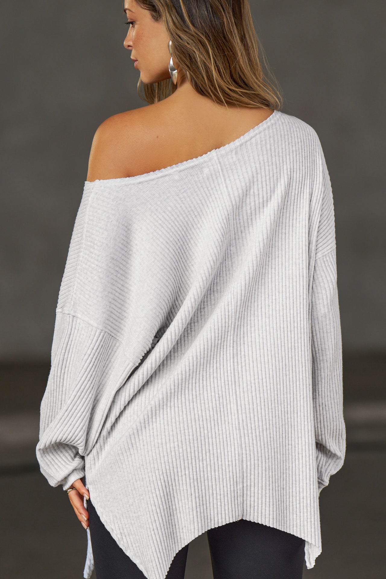 Back view of model posing in the relaxed fit pearl grey luxe knit Off Shoulder Tunic with a wide off-the-shoulder neckline and asymmetrical hem