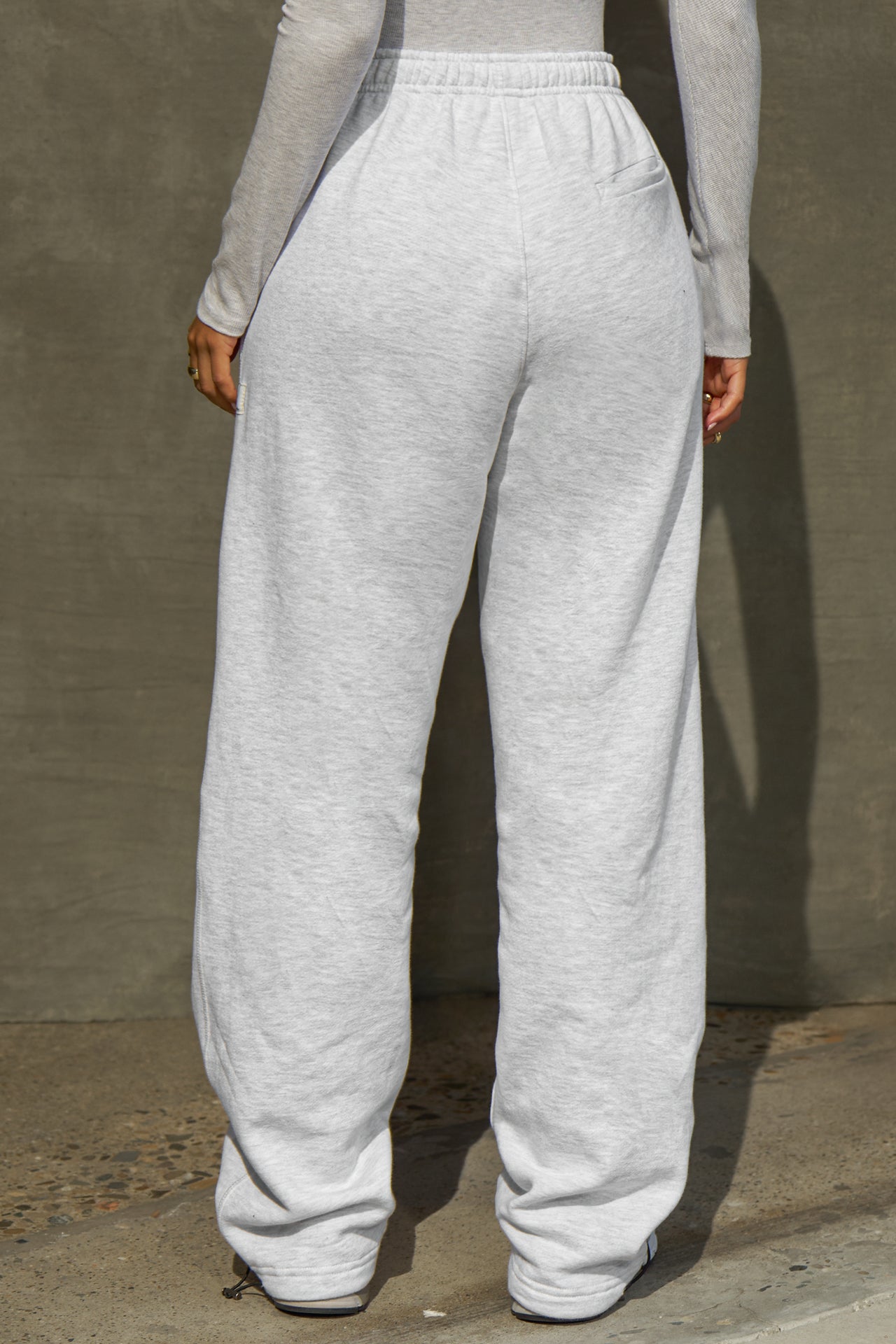 Back view of model from the waist down posing in the relaxed fit pearl grey french terry Oversized Straight Leg Jogger with an elastic waistband, hidden drawstring, pocket details. and adjustable cinching at the ankle cuffs