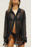 Front view of model posing in the sheer, lightweight black mesh Oversized Sheer Button Down long sleeve shirt with a classic collar, front button closures, dropped sleeves and buttons below the underarms for adjustable cinching.