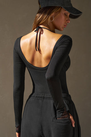 Back view of model posing in the fitted black flexrib Scoop Corset Long Sleeve with a low scoop back, adjustable ties at the neck, and thumbholes in the mesh sleeves
