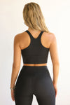 Back view of model posing in the fitted stretchy cropped black flexrib Varsity Tank Bra with a racerback silhouette