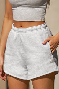 Close up front view of model from the waist down posing in the relaxed fit pearl grey french terry Classic Sweat Short with side pockets, hidden drawstring, adjustable elastic waistband and JOAH BROWN logo patch on the bottom left leg