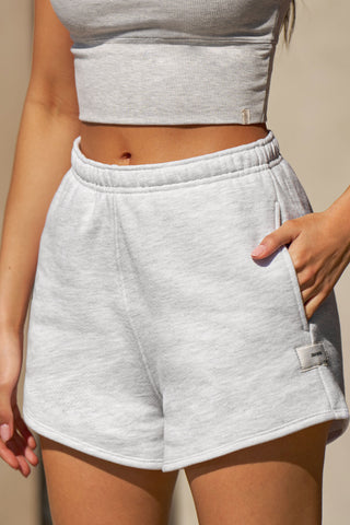 Close up front view of model from the waist down posing in the relaxed fit pearl grey french terry Classic Sweat Short with side pockets, hidden drawstring, adjustable elastic waistband and JOAH BROWN logo patch on the bottom left leg