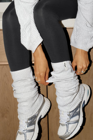 Close up detail view of model from the knees down adjusting the slouchy, pull on pearl grey luxe knit Leg Warmers that can be worn pulled up on scrunched down