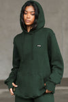 Front view of model posing in the oversized comfortable pine french terry with logo Empire Pullover Hoodie sweatshirt with a Joah Brown logo patch at the front left chest, kangaroo pocket and drawstrings at the bottom of the hood