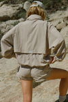 Back view of model posing outside in the relaxed-fit desert sage Classic Windbreaker Jacket with a padded collared neck for structure & adjustable corded details at the sweep for cinching