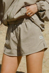 Close up front view of model from the waist down posing outside in the relaxed-fit desert sage nylon Classic Track Short with an elastic waistband, side seam pockets, and adjustable drawcord.