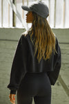 Back view of model posing in the loose fit cropped black french terry Cut Off Sweatshirt with oversized long sleeves, a raw cut hem and a white patch with a black joah brown logo on it sewn on the upper left chest