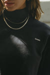 Close up detail front view of model posing in the loose fit cropped black french terry Cut Off Sweatshirt with oversized long sleeves, a raw cut hem and a white patch with a black joah brown logo on it sewn on the upper left chest