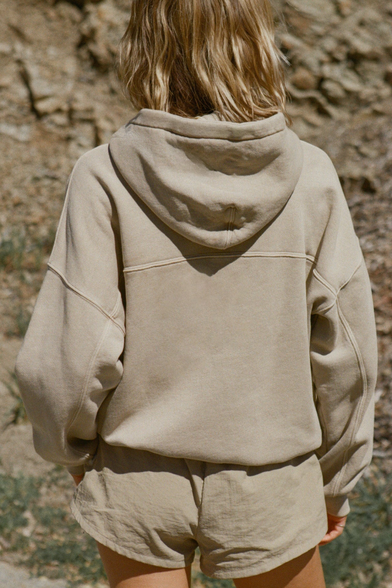 Back view of model posing outside in the oversized comfortable sand french terry Oversized Zip Hoodie with a full length front zip closure, side pockets and thumbholes in the fitted cuffs