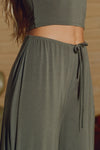 Close up detail side view of model from the waist down posing in the relaxed fit spruce modal Relaxed Straight Leg Pant with a wide leg, elastic waistband, adjustable drawstring tie and subtle pleating at the sides.