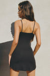 Back view of model posing in the form fitting stretchy black rib mini Slip Dress with thin straps, a u neckline and a tiny side slit at the hem