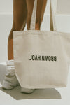 Back view of model holding the cotton canvas natural Tote Bag with shoulder straps and a black Joah Brown logo on the front