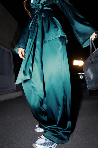 Close up front view of model from the bust down posing outside in front of a car in the soft and smooth classic pleated lagoon silk Relaxed Silk Trouser with a flattering wide leg, zip fly and side pockets.