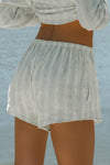 Close up back view of model from the waist down posing in the relaxed fit lightweight natural white and blue stripe gauze Relaxed Everyday Short with an elastic waistband and adjustable front tie