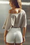 Back view of model posing in the relaxed fit and boxy sandstone cotton Off Shoulder Tee with a wide scoop neckline that can be worn off the shoulders.
