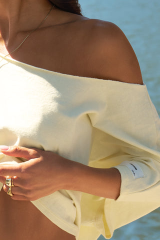 Close up detail front view of model posing outside by a lake in the relaxed fit and boxy buttercream cotton Off Shoulder Tee with a wide scoop neckline that can be worn off the shoulders.