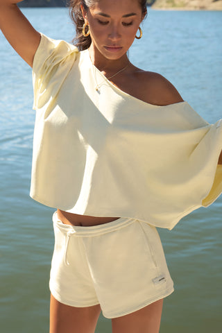 Front view of model posing outside by a lake in the relaxed fit and boxy buttercream cotton Off Shoulder Tee with a wide scoop neckline that can be worn off the shoulders.