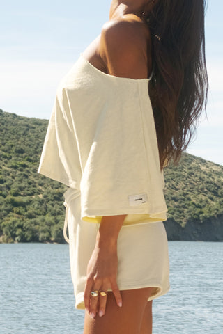 Side view of model posing outside by a lake in the relaxed fit and boxy buttercream cotton Off Shoulder Tee with a wide scoop neckline that can be worn off the shoulders.